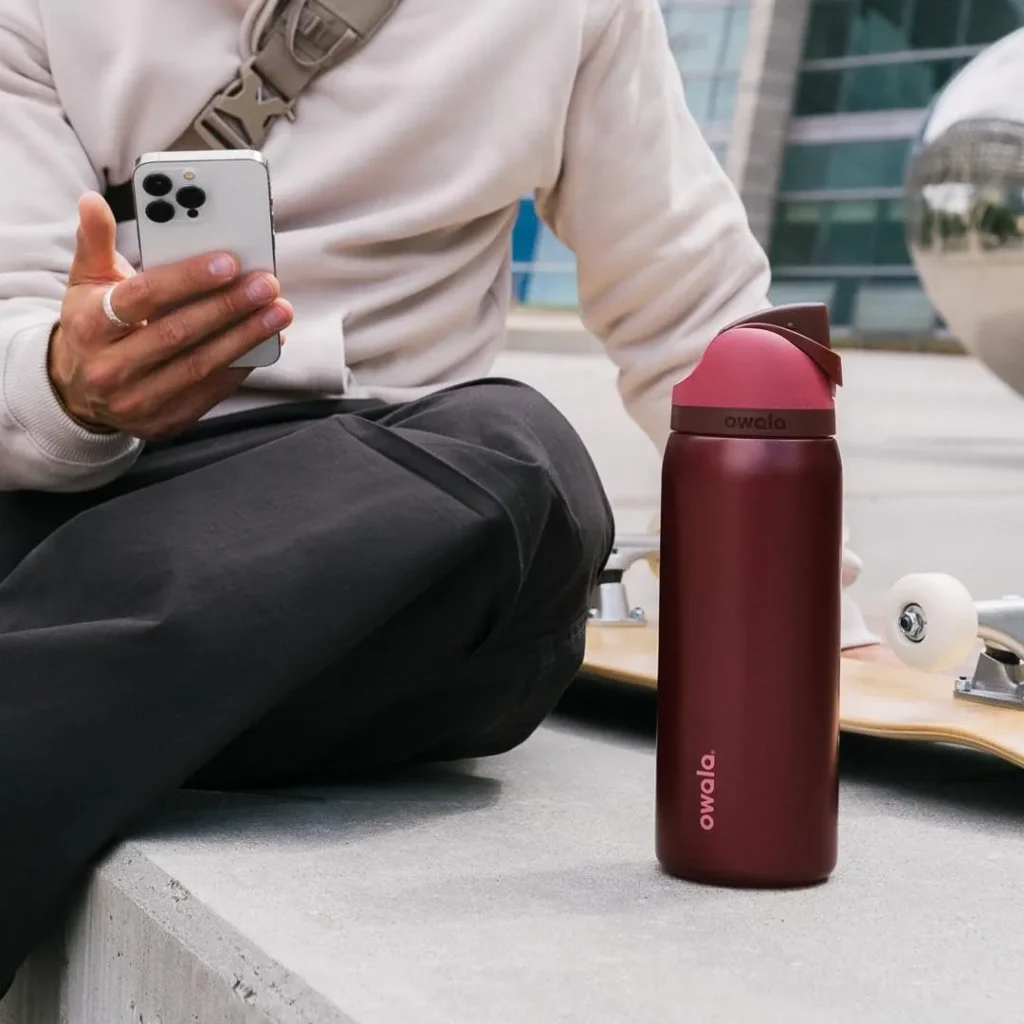 Person sitting outdoors with a skateboard, holding a smartphone next to a Black Cherry 32 oz insulated stainless steel water bottle with flip-top straw lid.