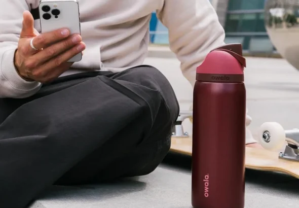 Person sitting outdoors with a skateboard, holding a smartphone next to a Black Cherry 32 oz insulated stainless steel water bottle with flip-top straw lid.
