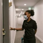 Woman wearing a face mask pressing a smart doorbell outside an apartment door in a hallway.