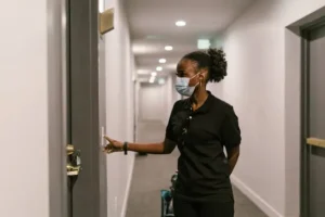 Woman wearing a face mask pressing a smart doorbell outside an apartment door in a hallway.