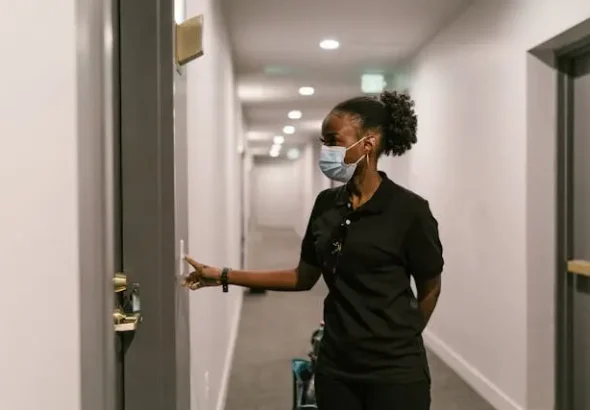 Woman wearing a face mask pressing a smart doorbell outside an apartment door in a hallway.