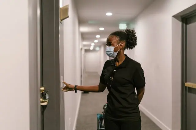 Woman wearing a face mask pressing a smart doorbell outside an apartment door in a hallway.