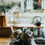 Kitchen sink area with drying dishes, mugs on a stand, knives in a block, and a teapot by a window.