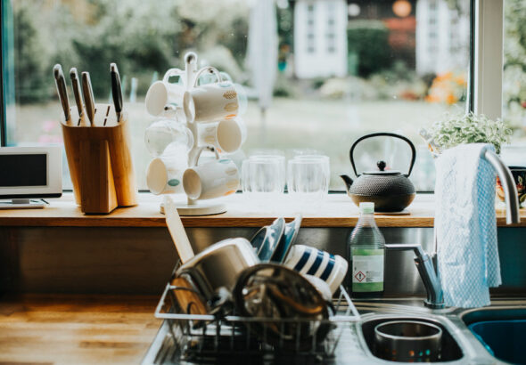 Kitchen sink area with drying dishes, mugs on a stand, knives in a block, and a teapot by a window.