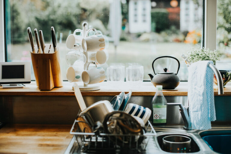 Kitchen sink area with drying dishes, mugs on a stand, knives in a block, and a teapot by a window.
