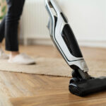 Person vacuuming a wooden floor with a cordless stick vacuum cleaner.