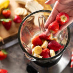 Hand adding fresh strawberries into a blender with sliced bananas.
