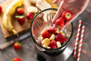 Hand adding fresh strawberries into a blender with sliced bananas.