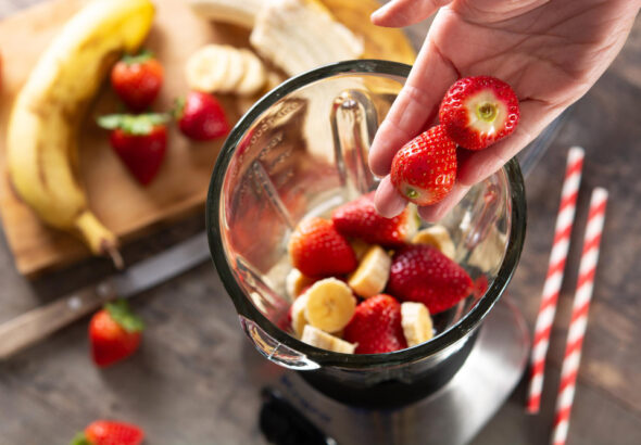 Hand adding fresh strawberries into a blender with sliced bananas.