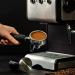 Hand holding a portafilter filled with freshly tamped coffee grounds next to a stainless steel espresso machine.