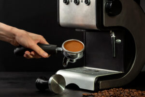 Hand holding a portafilter filled with freshly tamped coffee grounds next to a stainless steel espresso machine.