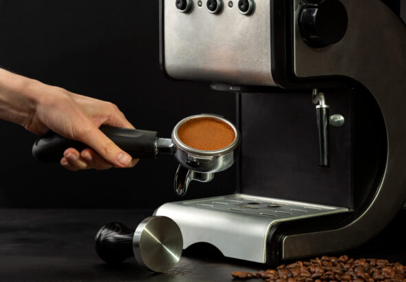 Hand holding a portafilter filled with freshly tamped coffee grounds next to a stainless steel espresso machine.