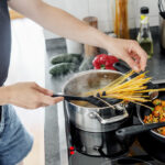 Person adding uncooked spaghetti into a pot of boiling water while vegetables cook in a pan nearby