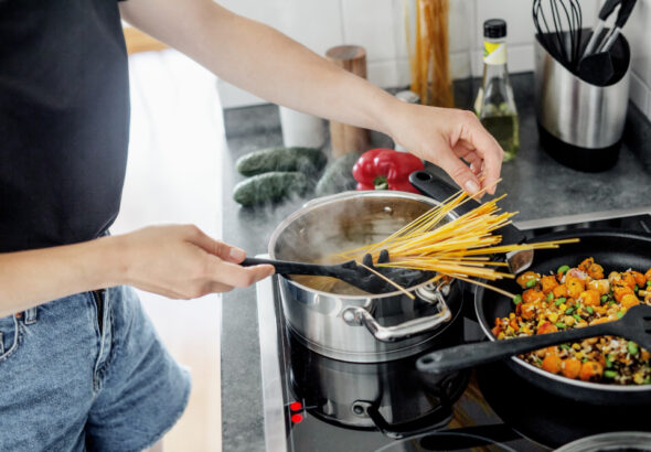 Person adding uncooked spaghetti into a pot of boiling water while vegetables cook in a pan nearby