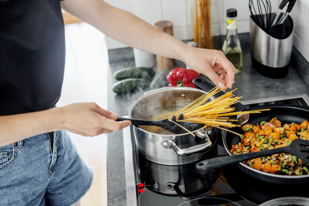 Person adding uncooked spaghetti into a pot of boiling water while vegetables cook in a pan nearby