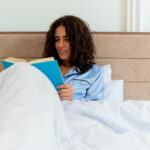 Woman in blue satin pajamas reading a book while relaxing on a bed with soft white bed sheets and pillows.