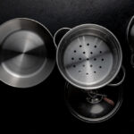 Top-down view of stainless steel cookware on a black surface, including a frying pan, a steamer insert, a pot lid, and a saucepan.
