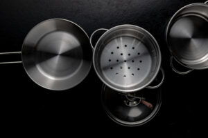 Top-down view of stainless steel cookware on a black surface, including a frying pan, a steamer insert, a pot lid, and a saucepan.