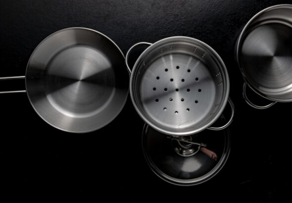 Top-down view of stainless steel cookware on a black surface, including a frying pan, a steamer insert, a pot lid, and a saucepan.