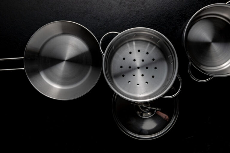Top-down view of stainless steel cookware on a black surface, including a frying pan, a steamer insert, a pot lid, and a saucepan.