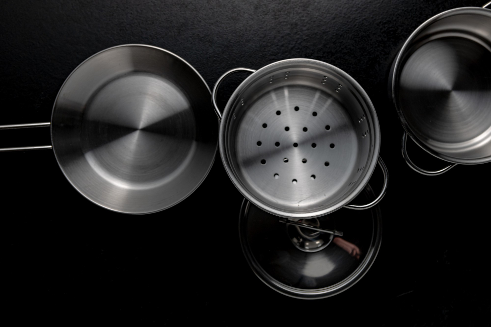 Top-down view of stainless steel cookware on a black surface, including a frying pan, a steamer insert, a pot lid, and a saucepan.