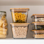Glass food storage containers with bamboo lids holding pasta, rice, chickpeas, and soybeans neatly organized on a pantry shelf.