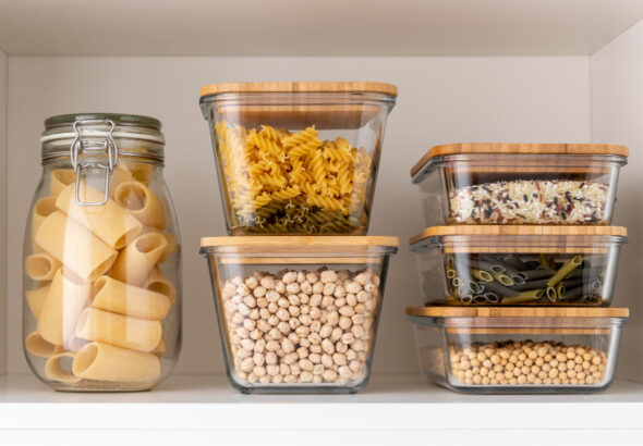 Glass food storage containers with bamboo lids holding pasta, rice, chickpeas, and soybeans neatly organized on a pantry shelf.