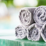 Soft gray bath towels neatly rolled and stacked on a wooden surface with a blurred spa-like background, creating a clean and relaxing atmosphere.