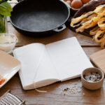 Cast iron skillet surrounded by fresh pasta, bacon, Parmesan cheese, eggs, basil, cream, and cooking ingredients on a rustic wooden table.