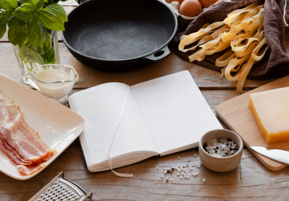 Cast iron skillet surrounded by fresh pasta, bacon, Parmesan cheese, eggs, basil, cream, and cooking ingredients on a rustic wooden table.