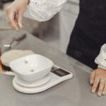 Person in a white eyelet blouse and dark apron carefully dropping small ingredients into a bowl placed on a digital kitchen scale on a stainless-steel countertop during baking preparation.