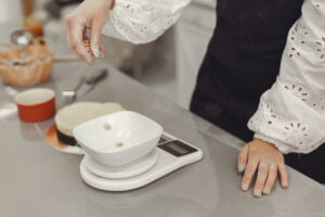 Person in a white eyelet blouse and dark apron carefully dropping small ingredients into a bowl placed on a digital kitchen scale on a stainless-steel countertop during baking preparation.