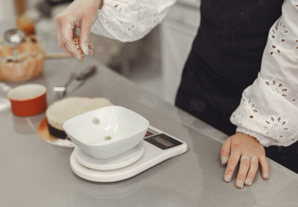 Person in a white eyelet blouse and dark apron carefully dropping small ingredients into a bowl placed on a digital kitchen scale on a stainless-steel countertop during baking preparation.
