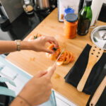 Person slicing tomatoes on a wooden cutting board with kitchen utensils, cooking oil, and sauce jars nearby in a home kitchen.
