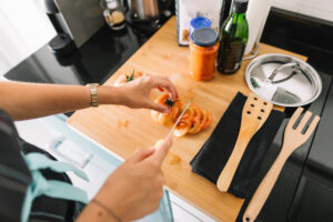 Person slicing tomatoes on a wooden cutting board with kitchen utensils, cooking oil, and sauce jars nearby in a home kitchen.