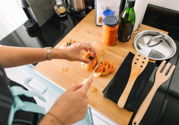 Person slicing tomatoes on a wooden cutting board with kitchen utensils, cooking oil, and sauce jars nearby in a home kitchen.